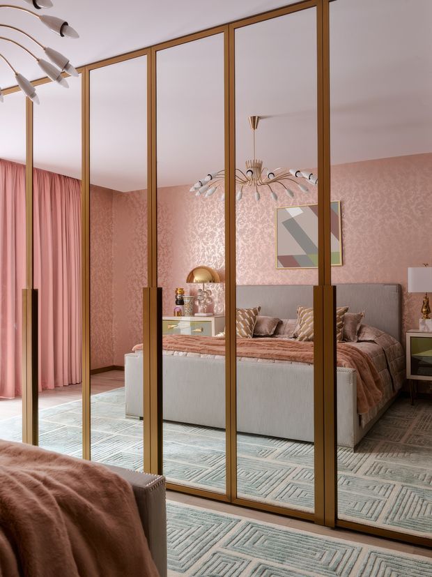 A richly styled bedroom viewed through full-height mirrored wardrobe doors with warm timber frames. The room features blush-pink patterned walls and curtains, a grey upholstered bed with dusty-rose bedding and cushions, geometric pale blue-grey carpeting, brass-toned accents, and sculptural mid-century-inspired pendant lighting. The mirrors reflect the bed and chandelier, making the space appear deeper and more layered.