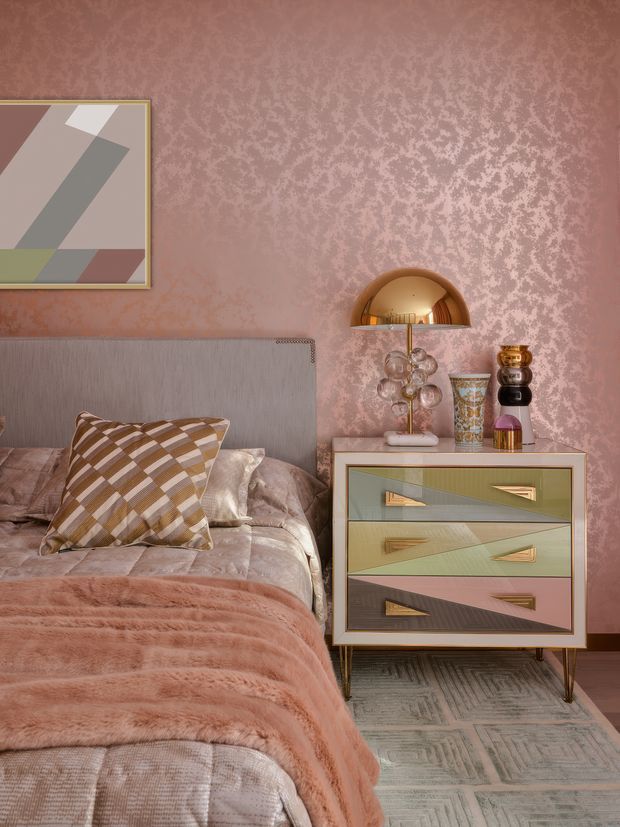 A richly styled bedroom vignette featuring a bed with a tall upholstered grey headboard, blush-pink textured wallpaper, layered mauve and taupe bedding, and a geometric patterned accent cushion. A colourful geometric-front bedside cabinet with tapered legs sits beside the bed, topped with a gold dome lamp, sculptural glass lamp base, decorative vessels, and a small patterned container. A framed abstract geometric artwork hangs above the headboard, while a pale patterned rug grounds the scene. The overall look is retro-inspired, glamorous, and highly coordinated in dusty pink, mauve, taupe, olive, cream, and metallic gold.