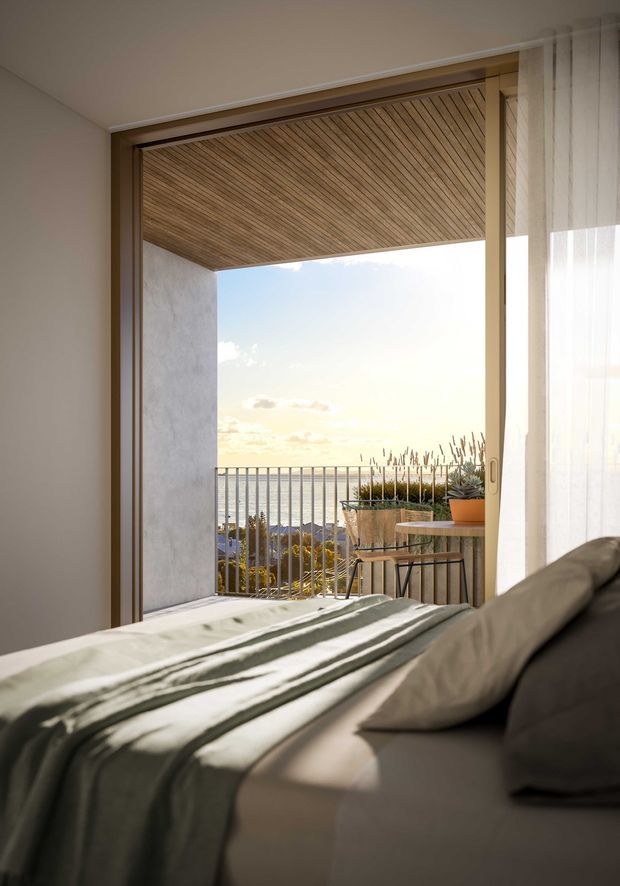 A calm, sunlit bedroom viewed toward a fully open glazed doorway leading to a balcony or terrace. The room has a neatly made bed with muted bedding, warm timber-framed glazing, sheer curtains, and a timber-lined soffit over the exterior opening. A simple outdoor table and potted plants sit beyond the balustrade, with a bright coastal horizon visible in the distance.
