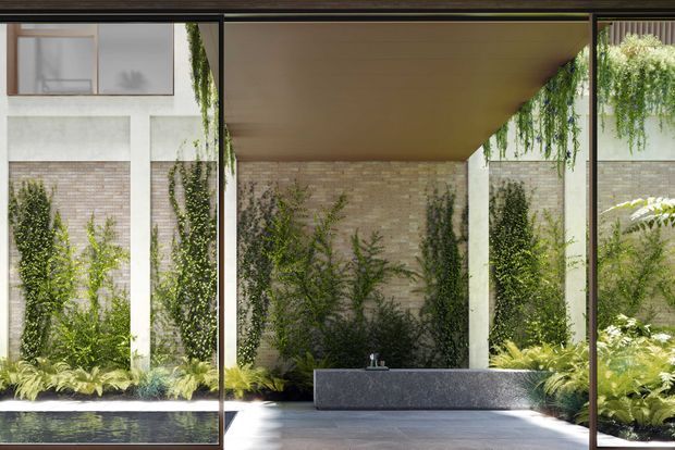 View from inside a glazed modern space toward a covered landscaped courtyard. Full-height dark-framed glazing surrounds a paved terrace, a long stone-clad raised planter or bench, and dense vertical greenery against pale brick and concrete walls. A warm timber-lined soffit extends over the courtyard, creating a calm indoor-outdoor transition.