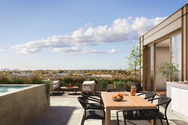A contemporary rooftop terrace or balcony with a tiled outdoor dining area, timber dining table, black woven chairs, a raised pool or water feature at left, planted screening, and large glazed openings to the building. The terrace overlooks a broad suburban cityscape under a bright sky.