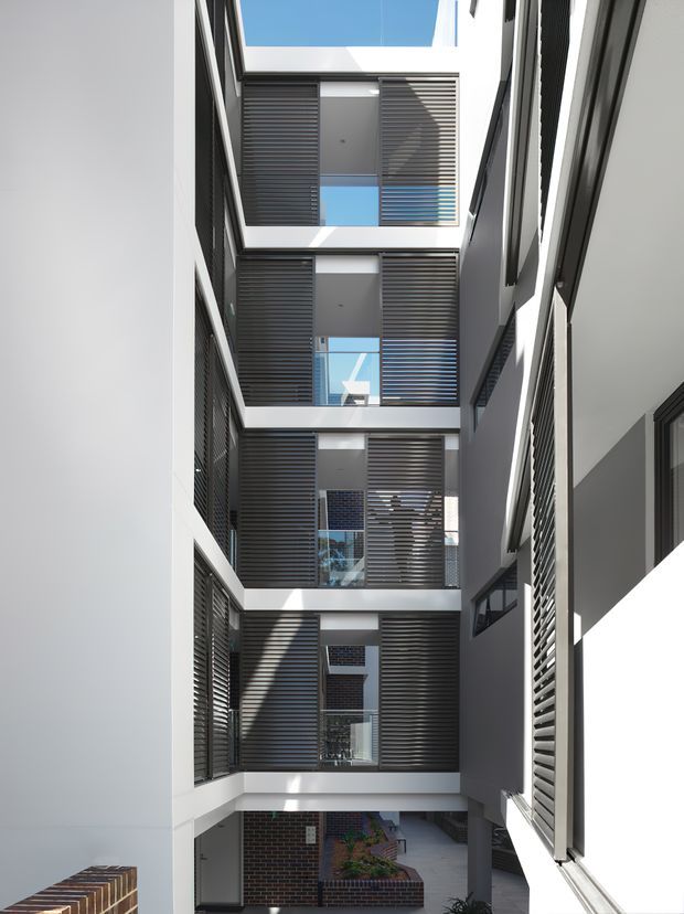 Vertical view through a multi-storey residential or commercial courtyard atrium, showing stacked open-air levels with extensive dark horizontal louvre screens, white and grey rendered walls, exposed balconies or walkways, and strong daylight from a roof opening. Brickwork and planted courtyard elements are visible at the lower level.