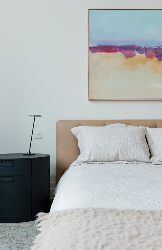 A softly styled bedroom with a tan upholstered bed, white textured bedding, a black curved bedside cabinet, and a slim black table lamp. A large abstract artwork in pale blue, cream, and muted burgundy hangs above the bed. The room has a calm, contemporary appearance with pale walls and a light textured rug or throw in the foreground.