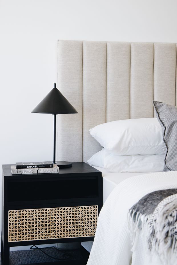 A close-up view of a refined bedroom featuring a tall vertically channelled upholstered headboard in light beige, crisp white bedding, and a black bedside table with a woven cane-front drawer. A matte black conical table lamp, stacked books, and monochrome cushions add contrast, while a textured grey-and-white throw rests across the bed.