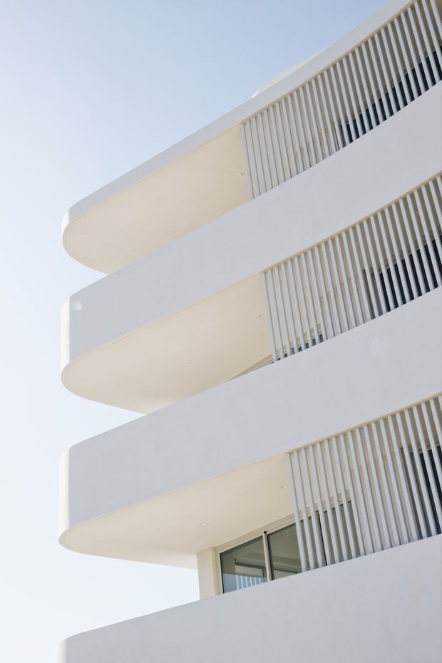 Upward-looking architectural photograph of a contemporary multi-storey building facade with stacked, rounded balcony slabs, white rendered surfaces, and repeated vertical privacy screens or balustrades. A small glazed opening is visible on the lower level against a pale blue sky.