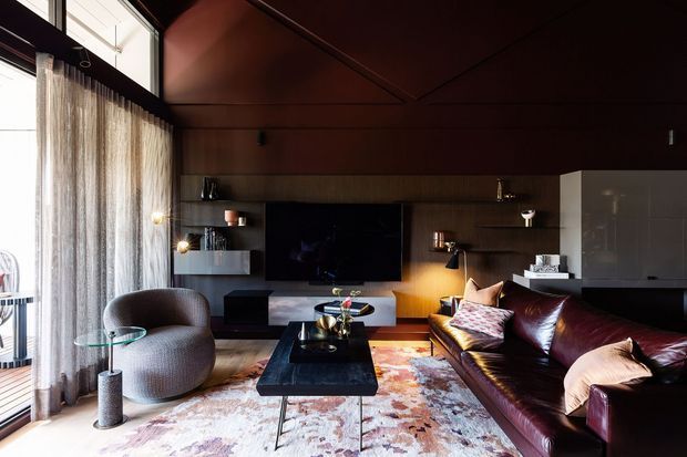 A sophisticated, moody living room with dark timber wall and ceiling panelling, floor-to-ceiling sheer curtains, a large television set into built-in joinery, sculptural armchair, black coffee table, patterned rug, and deep burgundy leather sofa. Warm accent lighting and a compact kitchen or joinery zone are visible at the rear.