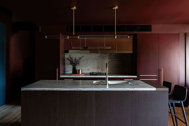 A dramatic, moody contemporary kitchen centered on a large island with a dark wood-grain base and pale stone benchtop. The kitchen features dark cabinetry, a textured stone backsplash, integrated appliances, a slim linear pendant light, brass-toned hardware and fittings, and a pair of counter stools. Deep burgundy and charcoal tones create a refined, high-contrast interior.