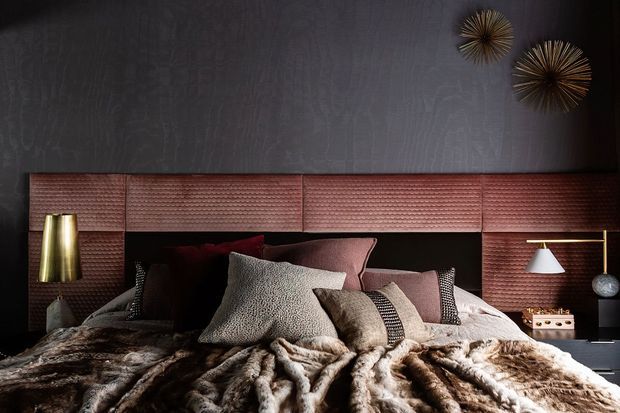 Moody, luxurious bedroom interior centered on a layered bed with a dark upholstered headboard, textured reddish-brown wall panelling, numerous neutral and burgundy cushions, and a plush fur-like throw. Brass and white bedside lamps, dark walls, and sculptural sunburst wall decor add refined warmth and contrast.