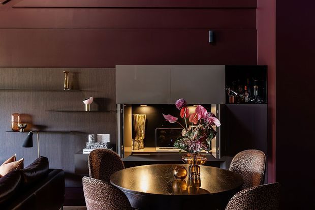A richly styled, moody dining and living area with a round dark dining table, textured upholstered dining chairs, floral arrangement, and a built-in entertainment and bar unit. Dark reddish-brown wall panelling, integrated shelving, warm metallic accents, and sculptural lighting create a refined, intimate atmosphere.