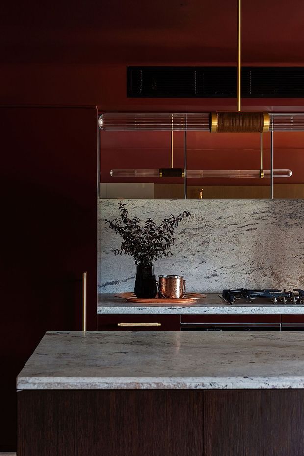 A dramatic, moody kitchen interior featuring a dark timber island with a pale veined stone benchtop, matching stone splashback, deep burgundy cabinetry and walls, brass hardware and accents, and a sculptural linear pendant light. A gas cooktop and integrated appliances are visible along the rear cabinetry, with a mirrored or reflective upper backsplash adding depth.