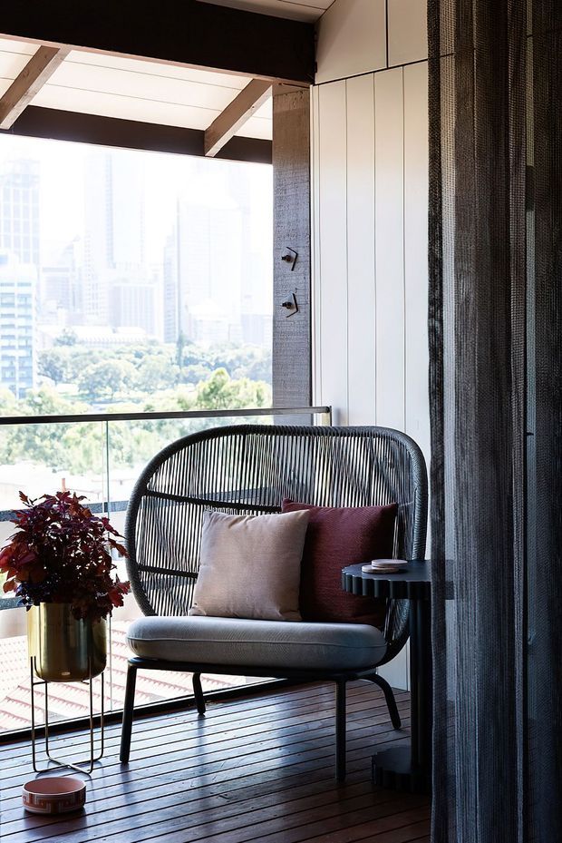 A bright, contemporary balcony or terrace seating area with an outdoor lounge chair, cushions, side table, potted plant, timber decking and a glass balustrade. The space is sheltered beneath a roof with exposed timber beams and framed by vertical white wall panelling and dark screened curtains, overlooking a leafy urban landscape.