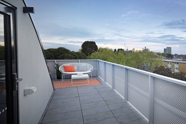 A rooftop or upper-level outdoor terrace with large grey tiled flooring, a light grey rendered wall, and white perforated metal balustrades. A compact white outdoor lounge set with a bright coral cushion sits on a terracotta-toned outdoor rug. The terrace overlooks leafy suburban and urban surroundings under an open sky.