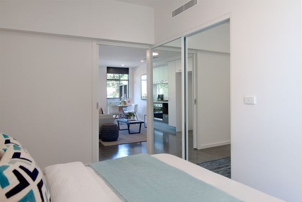 Bright, contemporary bedroom viewed toward a glazed sliding partition and adjoining open-plan living and kitchen area. The room has white walls, a pale blue and white bed covering, timber flooring, and mirrored or reflective glass wardrobe/partition doors. A dining area, kitchen cabinetry, and living furnishings are visible beyond the bedroom.