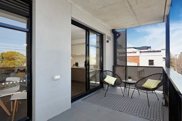 A modern apartment balcony or terrace with a raw concrete soffit and grey floor, enclosed by black metal-framed glazing and mesh balustrades. A compact black outdoor seating set with patterned cushions and a geometric outdoor rug creates a casual sitting area. The open glazed door reveals part of a contemporary kitchen and dining area inside.