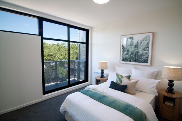 Bright, contemporary bedroom with a neatly made bed, white bedding, teal textured bed runner, coordinating cushions, matching timber bedside tables, and table lamps. A large black-framed window and glazed balcony door provide views of trees and an urban high-rise beyond. The room has white walls, a dark carpeted floor, and botanical artwork above the bed.