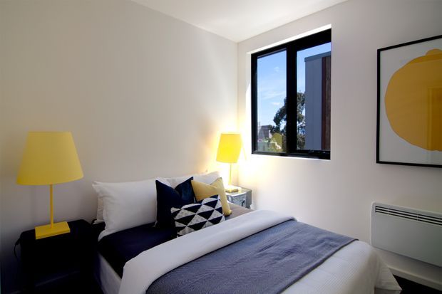 A compact, contemporary bedroom with a neatly made bed, white bedding, a dark navy throw, and geometric navy-and-white accent cushions. Two yellow lamps provide a bright colour accent beside the bed, while a black-framed window and minimalist abstract artwork complete the clean interior. A wall-mounted heater is visible beneath the artwork.