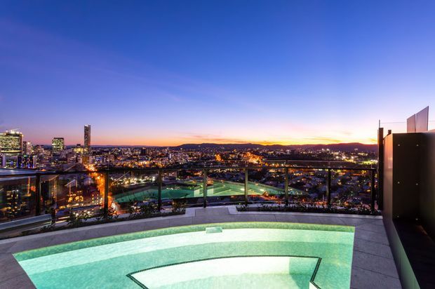 Twilight rooftop terrace overlooking a brightly lit urban skyline, with a glass balustrade, built-in illuminated swimming pool, and dark architectural privacy wall. The scene has panoramic city views and a sophisticated evening atmosphere.
