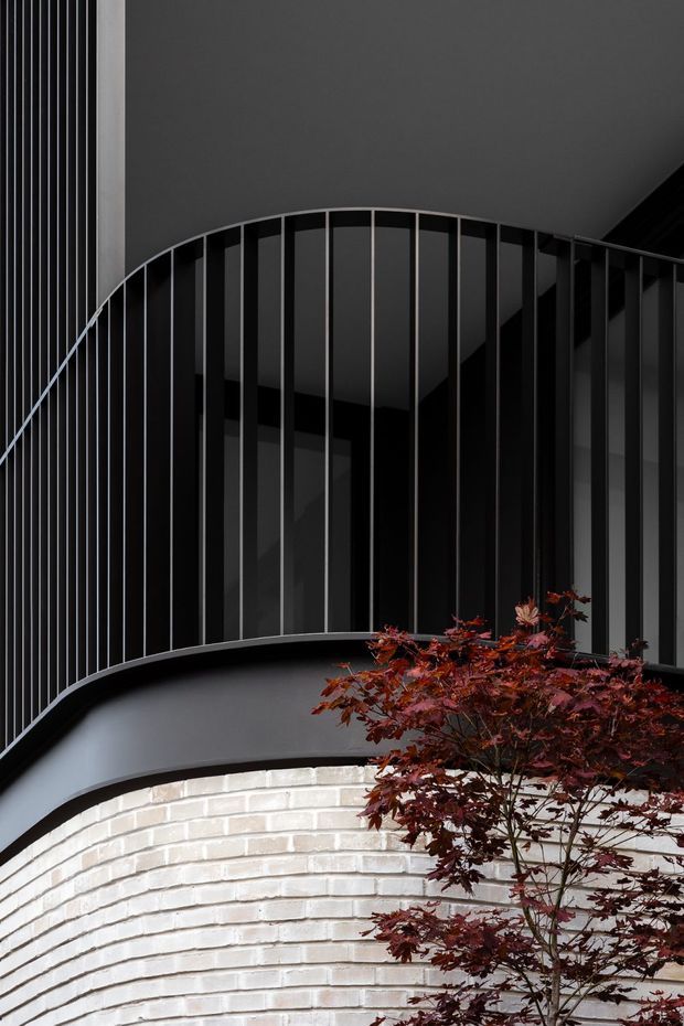 A close-up architectural view of a dark, sculptural curved stair or balcony structure. Slender black metal vertical balusters follow a sweeping arc above a curved black fascia, with a pale curved brick wall beneath. Deep charcoal surfaces and dramatic shadows create a refined, high-contrast composition, partially framed by a burgundy-leaved ornamental tree.