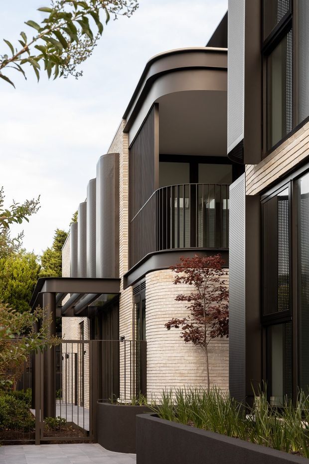 Contemporary multi-level residential facade with rounded projecting balconies, dark metal framing, perforated metal screening, and pale curved brickwork. A landscaped pedestrian entry path runs alongside dark raised planters, with slender trees and grasses softening the strongly geometric architecture.