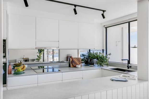 Bright, contemporary white kitchen with a large island, extensive handleless cabinetry, pale stone benchtops, integrated cooktop, black tapware, and expansive windows. Potted plants and timber cutting boards add natural accents, while black track lighting provides contrast.