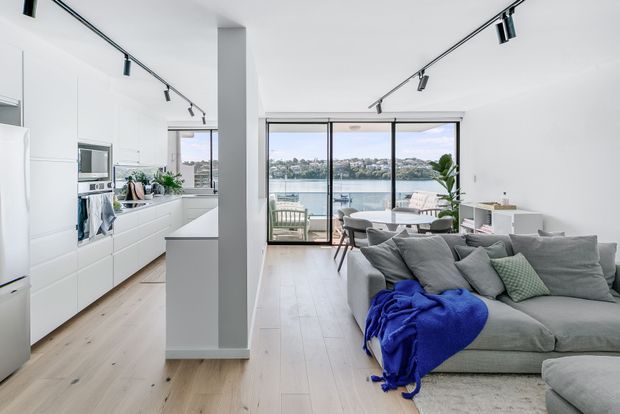 Bright open-plan contemporary apartment interior combining a streamlined white kitchen with integrated appliances and island, dining area, and a spacious grey sectional living room. Black track lighting and slim black-framed floor-to-ceiling glazing provide contrast, with a balcony and waterfront view beyond. Pale timber flooring and abundant natural light create an airy, modern atmosphere.
