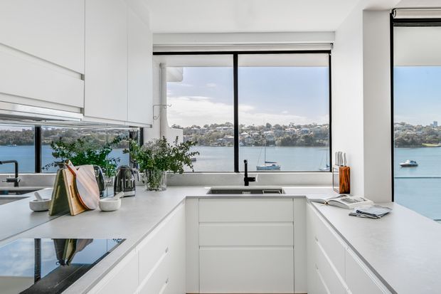 Bright contemporary white kitchen with an L-shaped peninsula and integrated cabinetry, positioned beside large black-framed windows overlooking a calm waterfront. The room features white benchtops and joinery, a black mixer tap, stainless-steel sink, induction cooktop, and pendant-free minimalist detailing.
