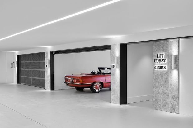 A clean, monochromatic modern parking garage interior with wide concrete floors, illuminated linear ceiling lighting, dark-framed garage openings, and a vintage red convertible parked in one bay. A textured stone-clad column carries signage for the lift lobby and stairs.