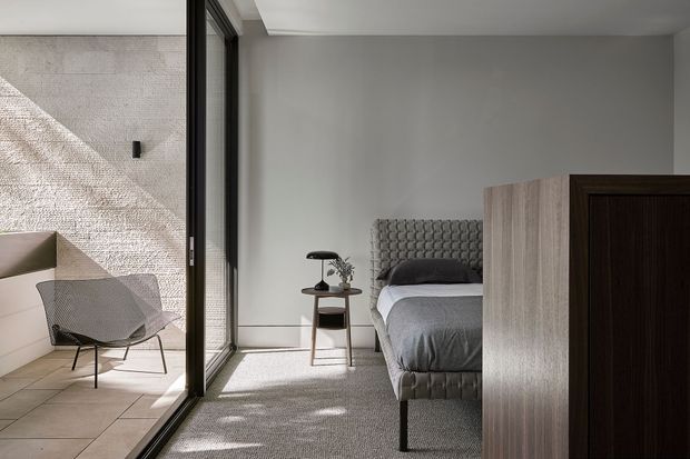 A contemporary bedroom opening onto a tiled outdoor terrace through expansive black-framed glazing. The room features a textured upholstered bedhead, grey bedding, a small timber-and-black bedside table, carpeted flooring, and a large timber-toned wardrobe or built-in cabinet. Strong sunlight creates patterned shadows across the neutral interior and adjacent terrace.