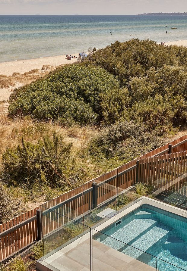 Elevated view of a tiled swimming pool and paved terrace enclosed by clear glass balustrades and timber screening, set behind native coastal vegetation with a sandy beach and calm blue sea beyond.