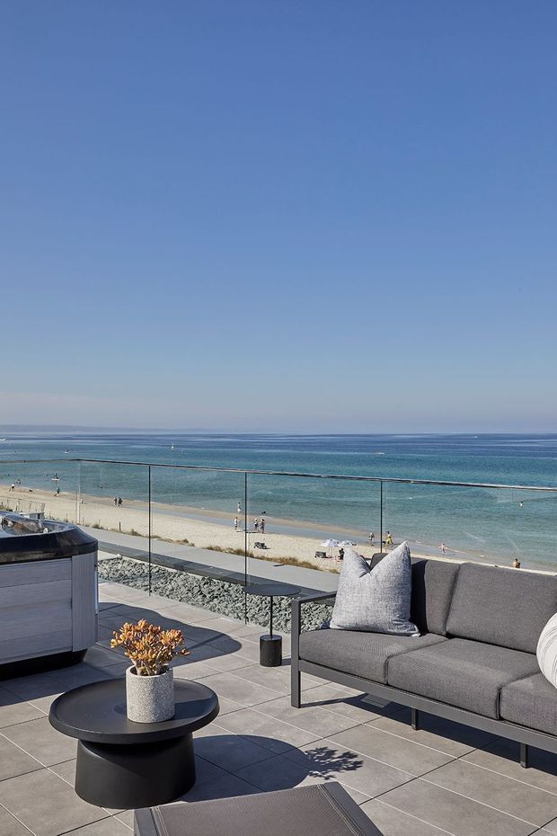 A sunny beachfront outdoor terrace overlooking a wide sandy beach and calm blue ocean. The terrace has large grey tiled paving, a frameless glass balustrade, a contemporary dark outdoor sofa with cushions, a round black coffee table, and a built-in outdoor cabinet or barbecue unit. The scene is bright, open, and coastal.