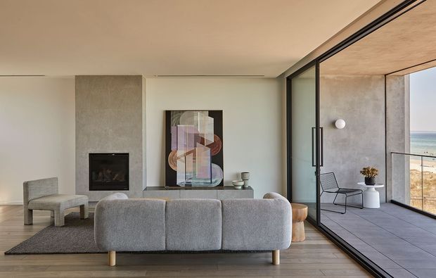 A refined contemporary living room with a low grey sectional sofa facing a textured fireplace wall and abstract artwork. Full-height black-framed glazing opens onto a tiled balcony or terrace with ocean views, while a sculptural lounge chair, side tables, and soft neutral finishes create a calm, minimalist setting.