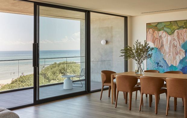 A refined dining area with a large timber dining table and upholstered brown dining chairs beside full-height black-framed sliding glass doors. The glazing opens to a balcony or terrace overlooking a sandy beach and ocean. A colourful abstract artwork, sculptural wall light, and vase with branches add decorative accents. The interior has a warm, contemporary coastal feel with abundant natural light.