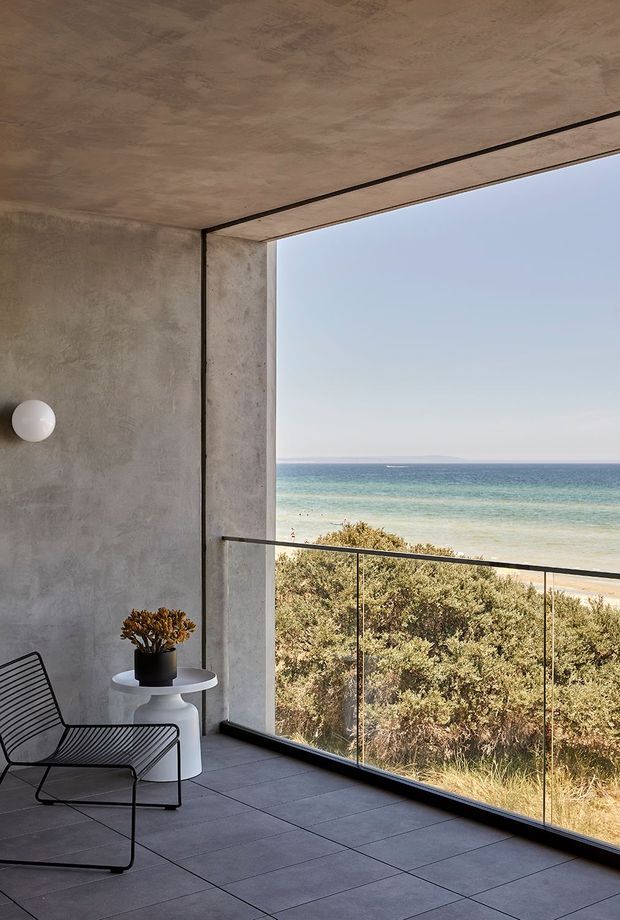 A minimalist coastal interior corner with a full-height glazed opening overlooking the beach and ocean. The space has raw textured concrete walls and ceiling, large dark grey tiled flooring, frameless-looking glass balustrading, a black wire lounge chair, a small white pedestal side table, and a round wall light. Natural light and the uninterrupted sea view dominate the scene.