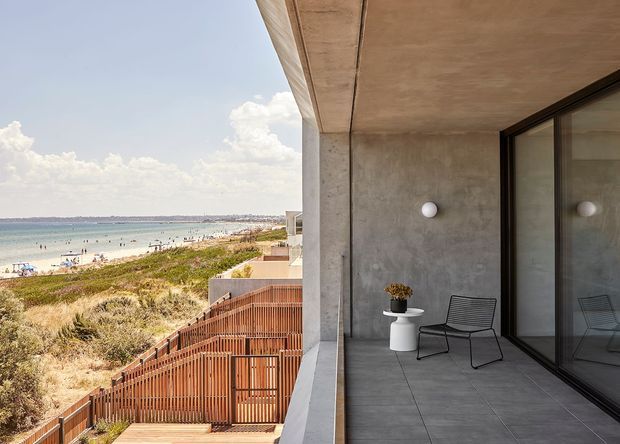 A minimalist concrete balcony or terrace overlooking a sandy coastal beach and ocean. The space features a smooth grey concrete wall and soffit, large dark-framed sliding glass doors, grey tiled flooring, a small white side table with a potted plant, and a black wire-frame chair. Timber privacy fencing and dune vegetation extend below the balcony.
