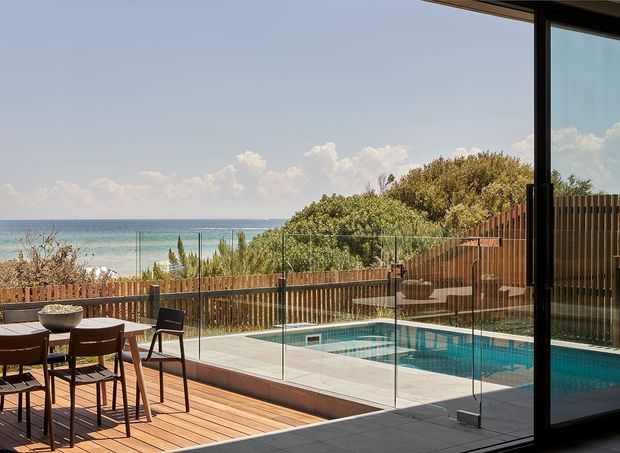 A contemporary coastal outdoor entertaining area viewed from inside through expansive black-framed glazing. A timber deck with a dining table and chairs overlooks a glass-balustraded swimming pool, timber privacy fencing, landscaped planting, and a clear ocean horizon beyond.