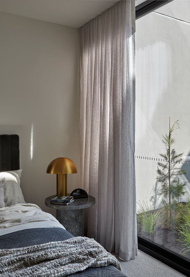 A calm, refined bedroom corner with a partially visible upholstered bed, layered grey and cream bedding, a dark stone round bedside table, and a sculptural brass dome lamp. Full-height sheer grey curtains frame large black-framed glazing overlooking a planted courtyard or garden, while soft natural light creates a muted, contemporary atmosphere.