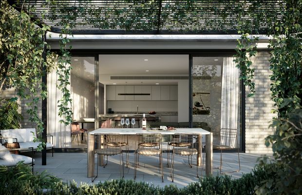 A refined outdoor dining terrace viewed toward a modern kitchen through wide open glazed doors. The scene features a long dining table with wire-frame chairs, a kitchen island beyond, pale masonry walls, large sliding glass openings, climbing greenery, and a slatted overhead pergola casting patterned shade.