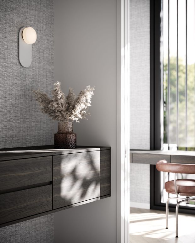 A refined contemporary interior vignette showing a dark wood floating sideboard with integrated drawers against a textured grey feature wall. A sculptural wall light and a ceramic vase with dried branches sit above the joinery. Through the adjacent opening, a bright dining area is visible with a timber-topped table, leather dining chairs, large black-framed windows, and strong natural light.