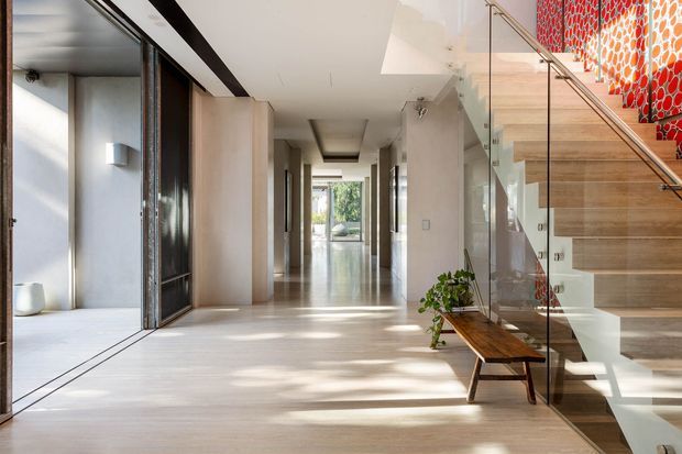 Bright contemporary interior hallway with pale timber flooring, full-height glazing, minimalist white walls, and a sculptural open staircase. A glass balustrade and timber stair treads lead toward a bold red-and-white patterned feature wall, with a narrow timber console and potted plant beneath the stairs.