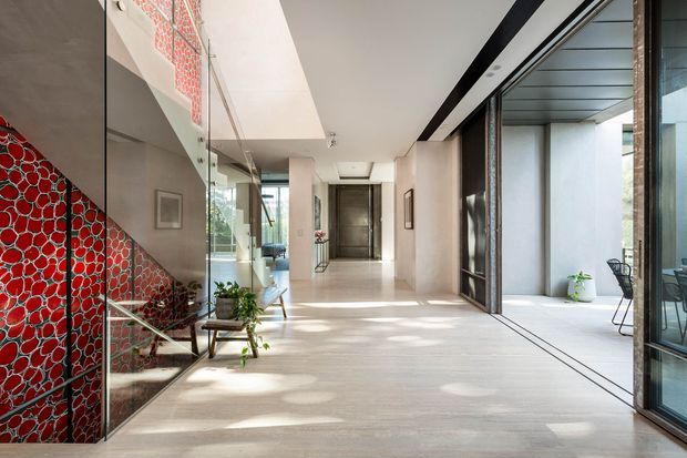 A bright, spacious contemporary hallway or entry corridor with pale stone or concrete flooring, white walls, and full-height glazing along one side. A dramatic red patterned feature wall and matching stair enclosure create a strong accent, while a glass balustrade, dark-framed internal doors, and minimalist furnishings reinforce the modern design. The corridor leads toward a dark entry door, with an outdoor courtyard or terrace visible through the glazing.