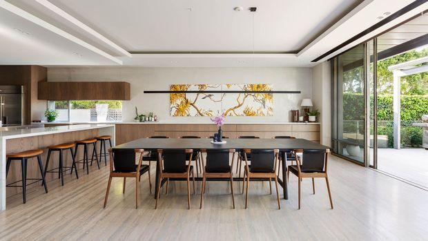 A spacious open-plan dining and kitchen area with a long dark dining table surrounded by wooden chairs, a linear kitchen with timber cabinetry and light benchtops, and bar seating along the island. Full-height glazed doors open to a landscaped outdoor area. Recessed ceiling detailing and a large botanical artwork add visual interest.