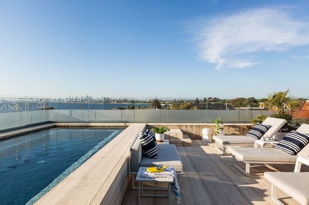 Rooftop terrace with a rectangular swimming pool, glass balustrades, timber-look decking, pale stone pool coping, and white outdoor lounge furniture with navy striped cushions. The elevated view overlooks a marina or waterfront and a distant urban skyline under a clear sky.