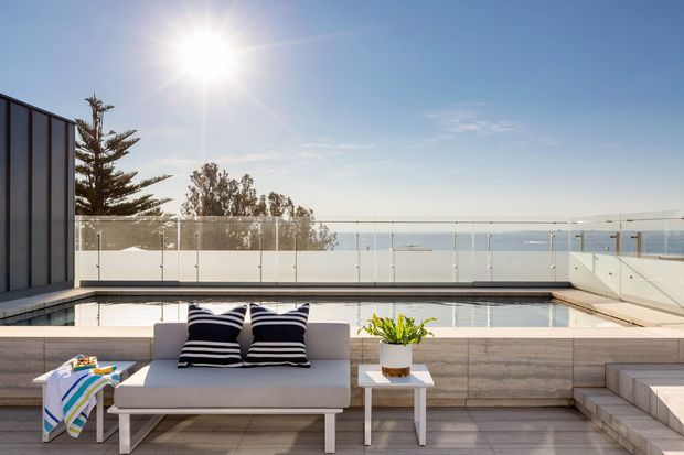 Sunlit rooftop terrace with a shallow reflecting or plunge pool, frameless glass balustrade, built-in tiled seating, and a simple outdoor sofa with striped cushions. The terrace overlooks a calm coastal or waterfront setting, with broad open sky and distant water visible beyond the railing.