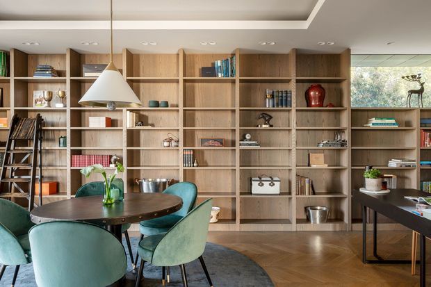 A refined open-plan dining and living area dominated by a full-height timber built-in shelving wall filled with books and decorative objects. A round dark dining table with teal upholstered chairs sits beneath a white pendant light, while a dark desk or console occupies the right side. The room has a recessed ceiling detail, warm timber flooring, neutral walls, and abundant natural light.