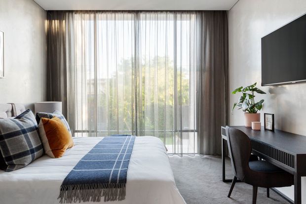 A refined contemporary bedroom with a large floor-to-ceiling glazed opening dressed in sheer curtains and dark drapes. The room features a neatly made bed with layered blue, grey, and mustard textiles, a dark streamlined desk and chair beneath a wall-mounted television, soft grey carpet, pale textured walls, and a potted plant. The space is bright, calm, and understated.
