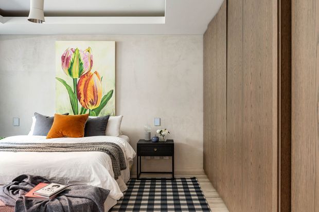 A warm, contemporary bedroom with a neatly made bed, layered neutral bedding, a rust-orange accent cushion, and a checked rug. A colourful floral artwork hangs above the bed, while a compact dark bedside table and extensive light timber built-in cabinetry line the room. The walls and ceiling are finished in soft neutral tones with recessed ceiling detailing and a small pendant light.