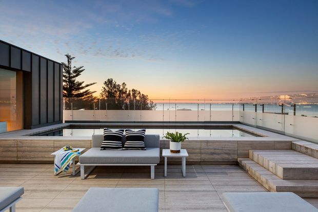 A contemporary rooftop terrace overlooking a coastal waterfront at sunset. The spacious terrace features pale large-format tiled paving, stepped built-in seating, a glass balustrade, a modern outdoor sofa with striped cushions, and a dark vertically panelled building facade at left.