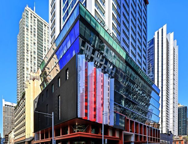 Exterior architectural photograph of a contemporary multi-storey urban building, with a dark lower volume, red structural framing, projecting translucent vertical fins, and a dramatic angled glass curtain wall. Dense high-rise towers surround the building under a clear blue sky.