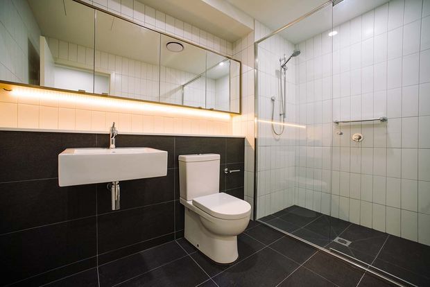 Contemporary bathroom with a wall-mounted white basin, toilet, and frameless glass shower enclosure. Large mirrors span the vanity wall, while white grid-pattern wall tiles contrast with dark charcoal wall and floor tiles. Warm concealed lighting beneath the mirrored cabinet adds a soft glow.