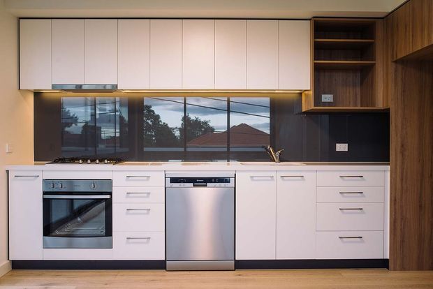 A compact contemporary kitchen with white flat-panel overhead and base cabinetry, integrated appliances, a dark reflective splashback, timber-toned side cabinetry, and warm under-cabinet lighting. The kitchen includes an oven, dishwasher, cooktop, sink, drawers, open shelving, and light timber flooring.
